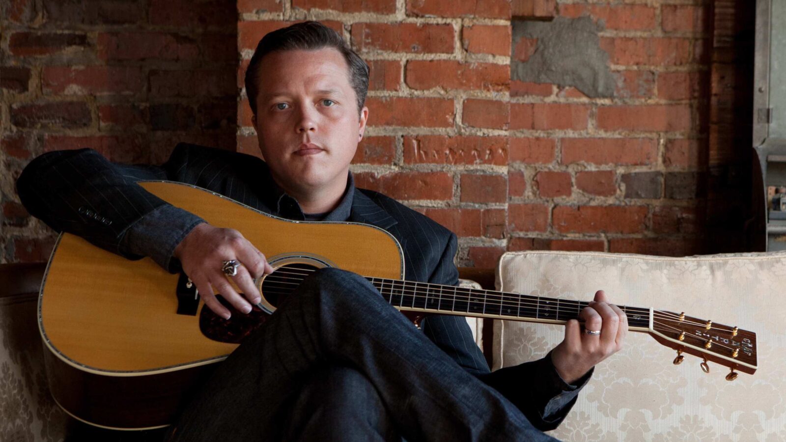 A man holds a guitar and sits on a couch