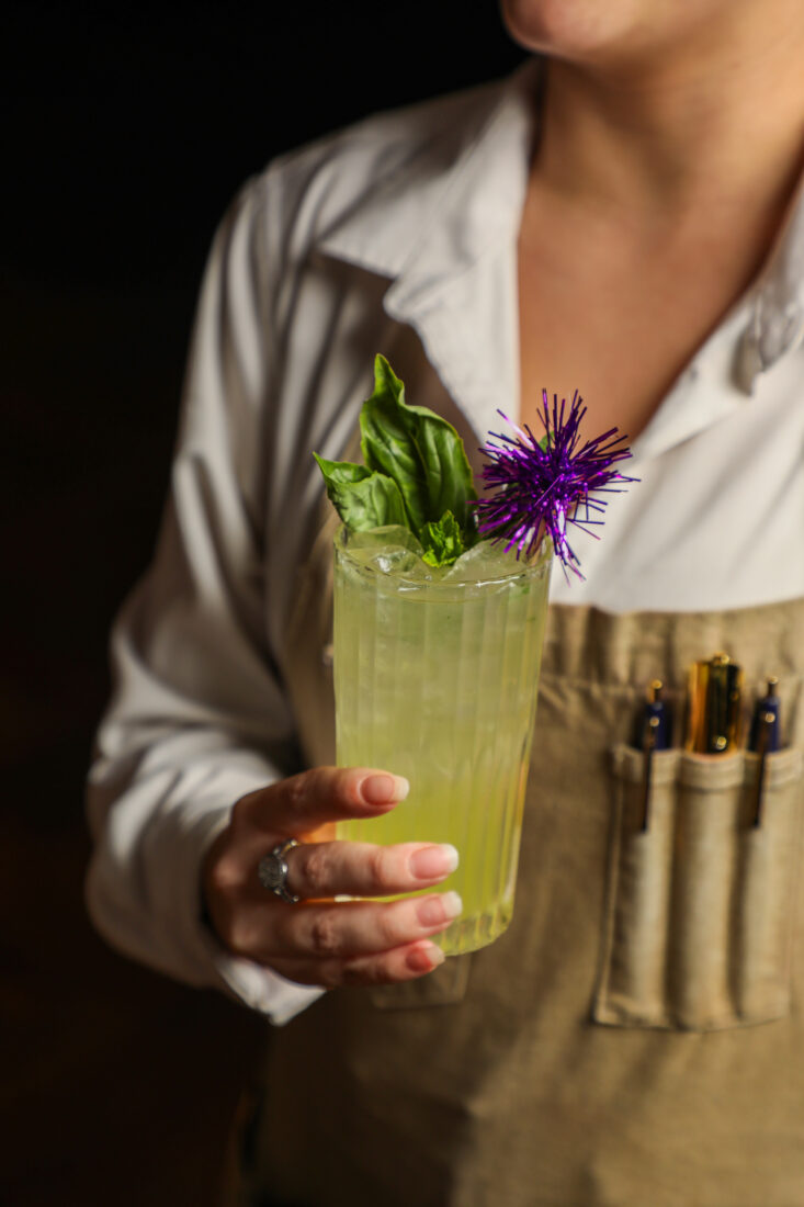 A person wearing a white shirt and beige apron holding a green drink with basil garnish and purple sparkler.