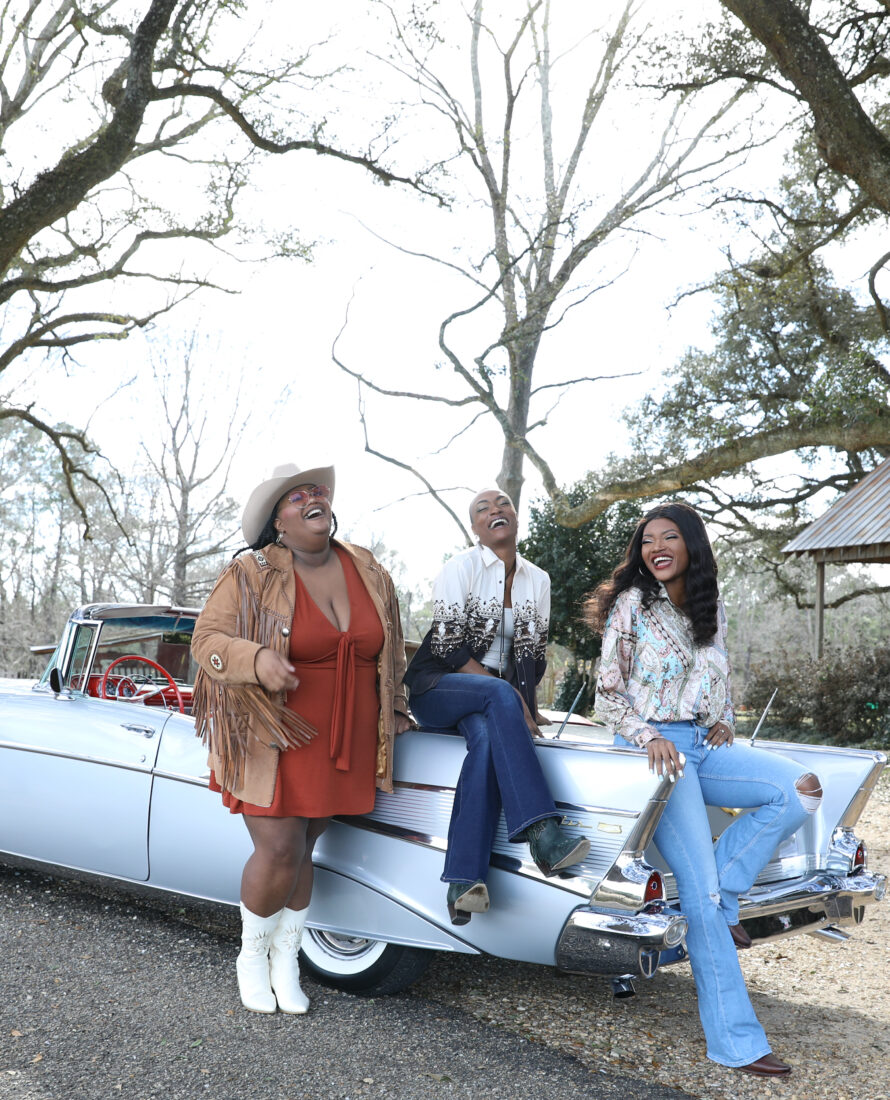 Three woman sit on a vintage car, laughing