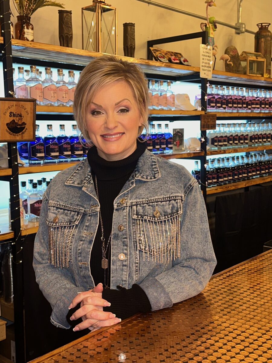 A woman stands behind a bar with bottles of moonshine behind her in the shelves