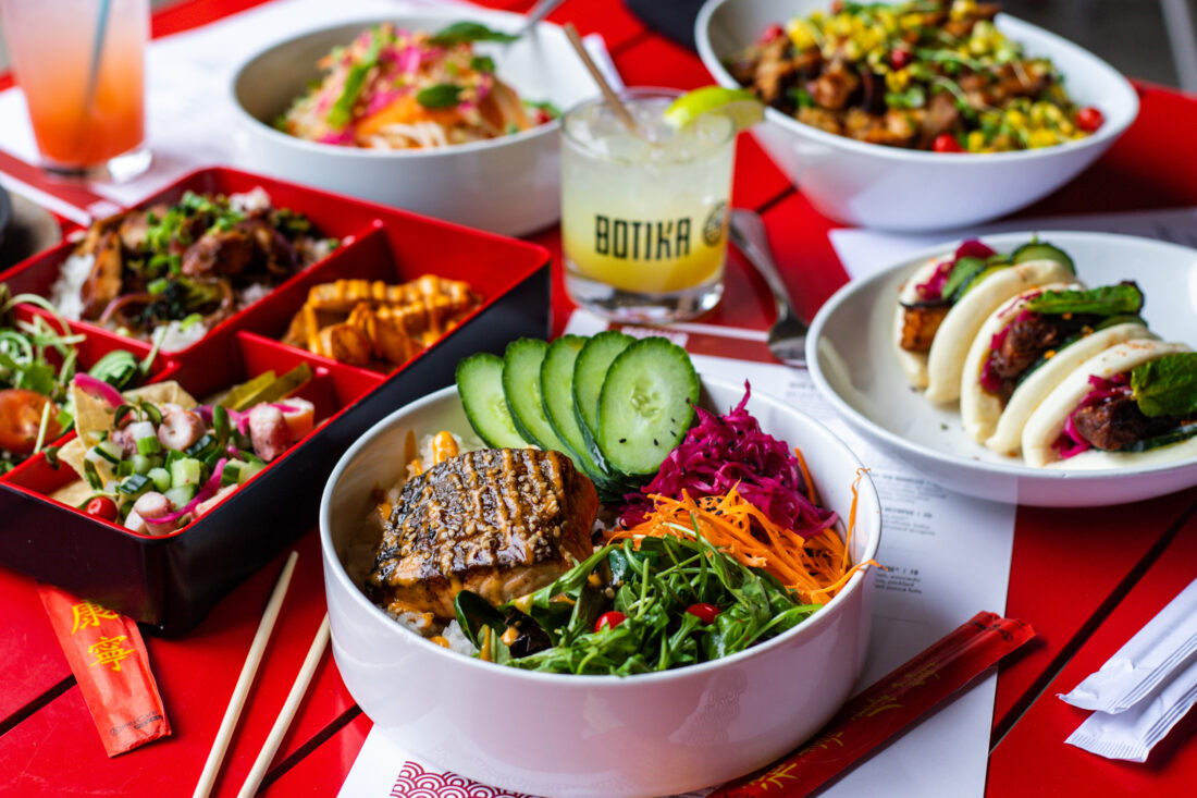 A spread of lunch food on a red table, including a bento box with rice and veggies, bao buns, and a bowl of seared fish with salad and rice