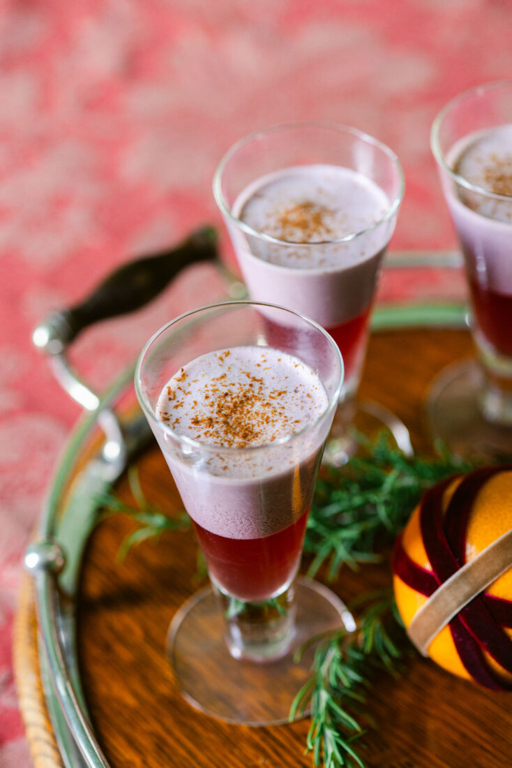 A glass of red wine with foam on top and spices on the foam; it is on a blurred background of a wood round bar cart and a red rug. There is pine greenery and an orange by the glass