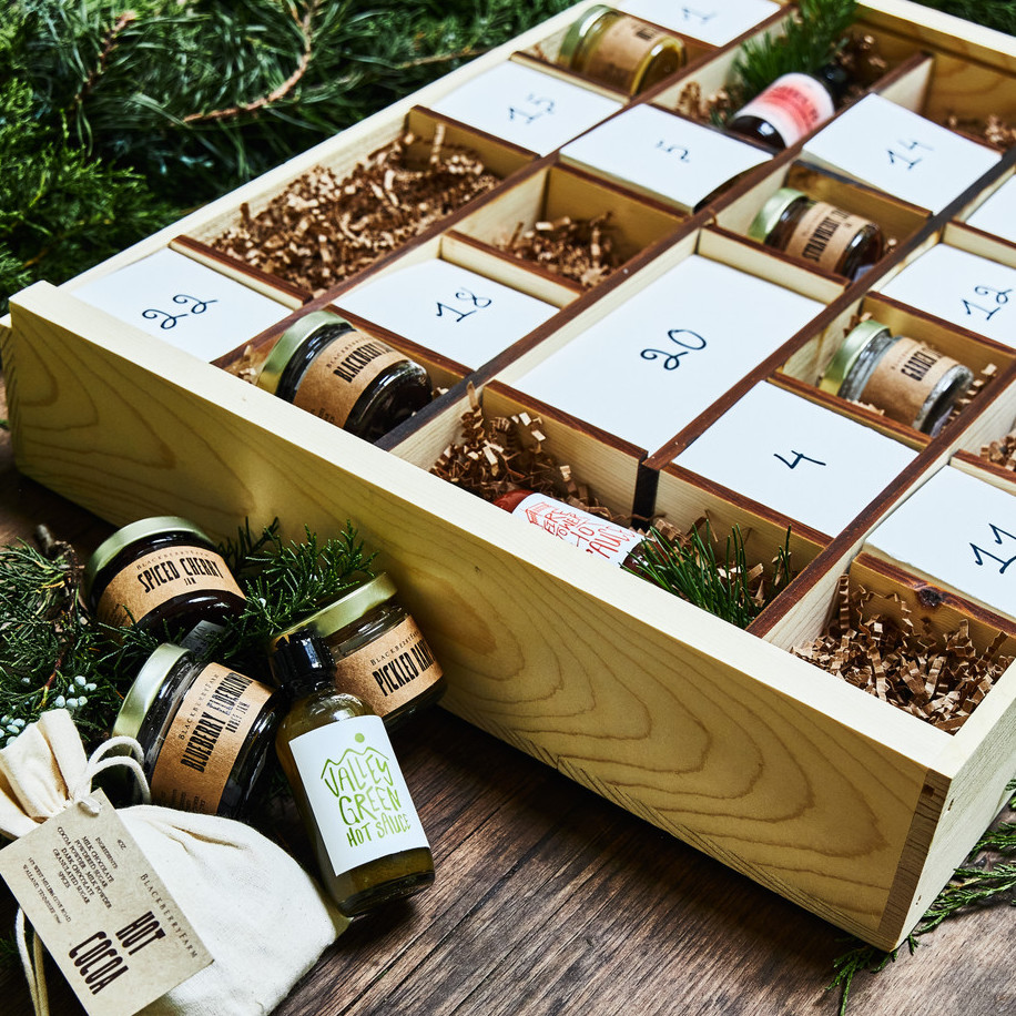 A wooden advent box with numbers; some are open to reveal jars of jam and sauces. Various jars of sauce, pickled veggies, and jams lay in the bottom left corner