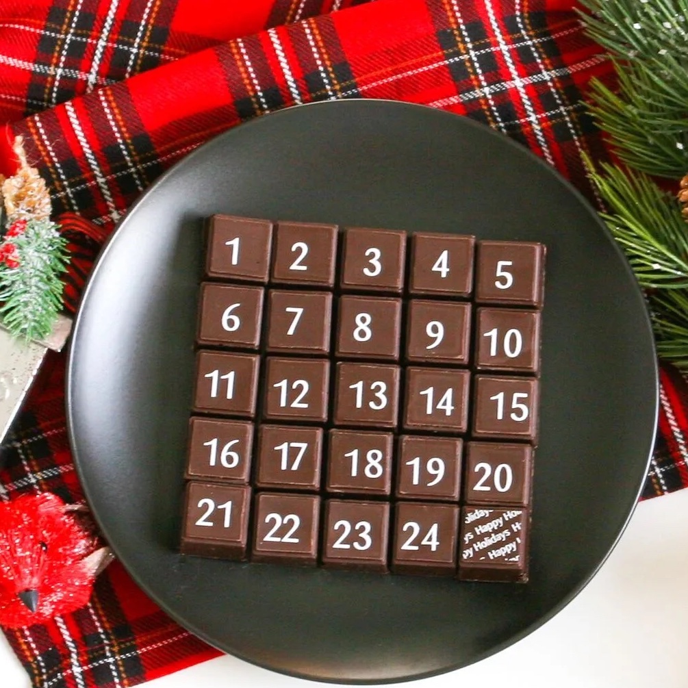 A square of chocolates (shaped into smaller squares, numbered 1-25) on a black plate. The plate lays on a red plaid background with greenery.