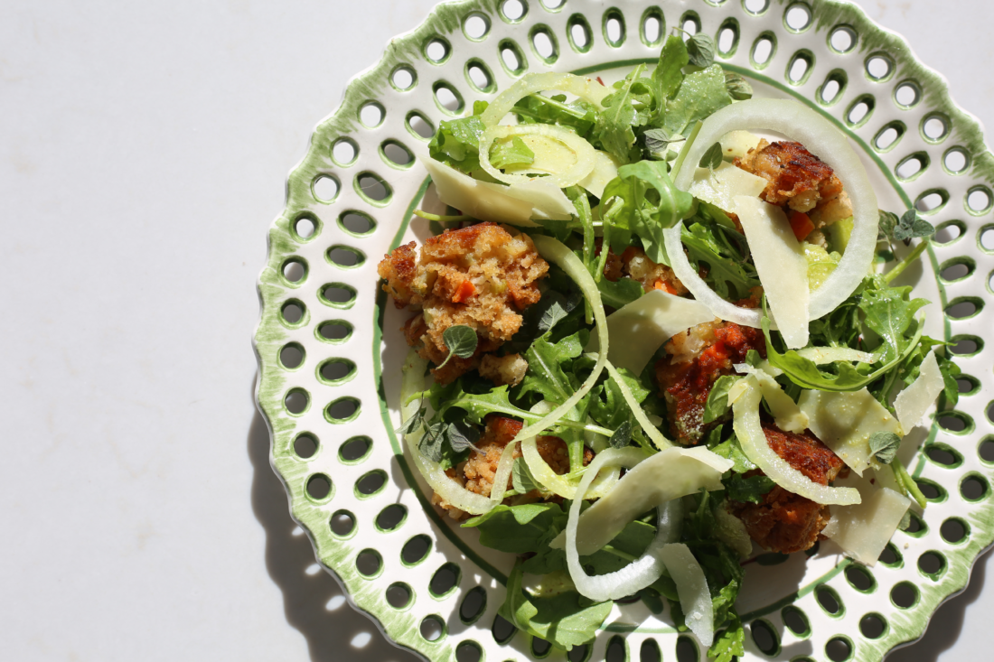 A green and white round plate holds salad greens, shaved white onion and cubes of golden brown stuffing.