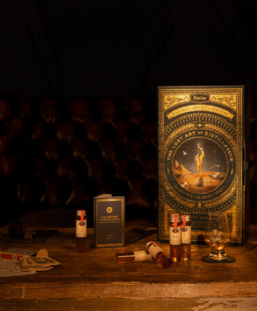 A gold advent calendar stands on a wood table in a dark room; it has whiskey samples, Glencairn glass, leather coaster, and a tasting journal.