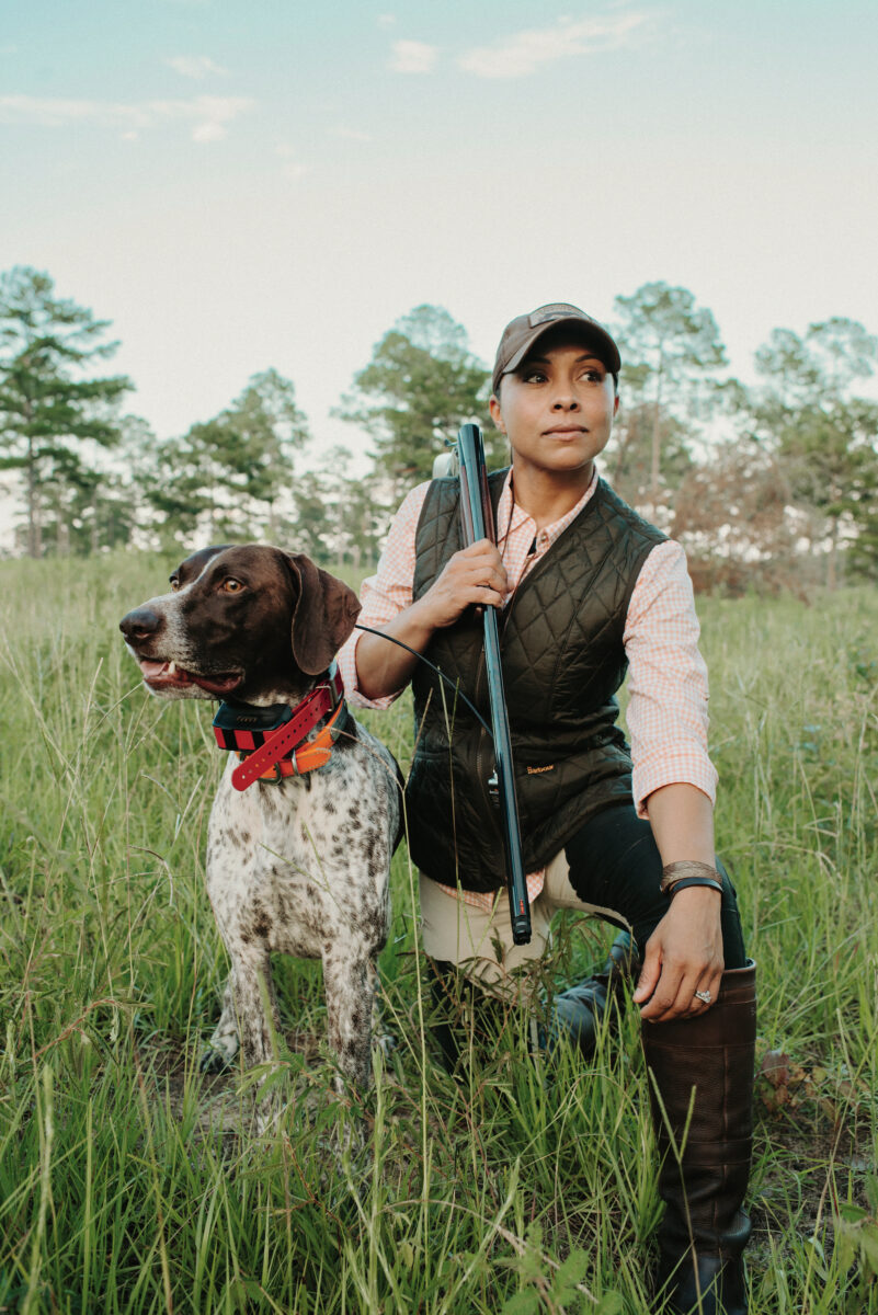 A woman kneels in a grassy field with her dog, a shorthaired pointer with white and brown fur, to the left