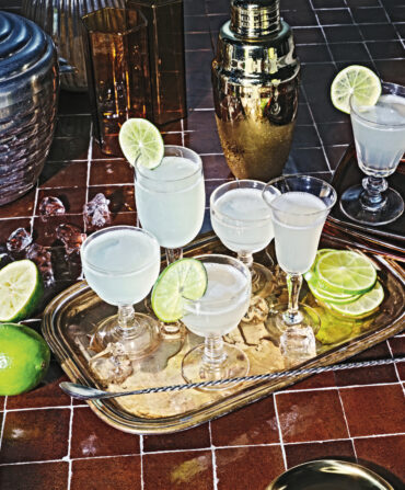 A gold platter of cocktails with lime garnishes and ice. The platter is on a red tile background with a sliced lime, shaker, ice, and glasses