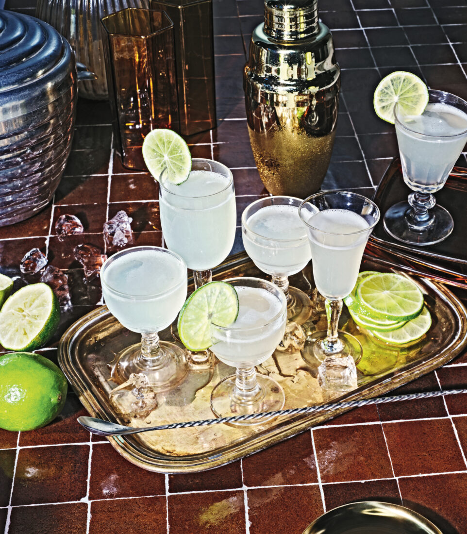 A gold platter of cocktails with lime garnishes and ice. The platter is on a red tile background with a sliced lime, shaker, ice, and glasses