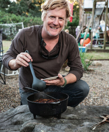A man crouches in a backyard with a cast-iron pot of cobbler