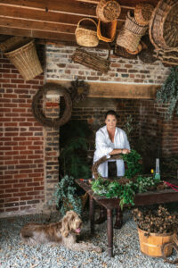 A woman stands behind a table with wreath-making supplies. There is a Spinone Italiano dog at her feet. A giant brick fireplace is behind her, and wicker baskets hang from the ceiling.
