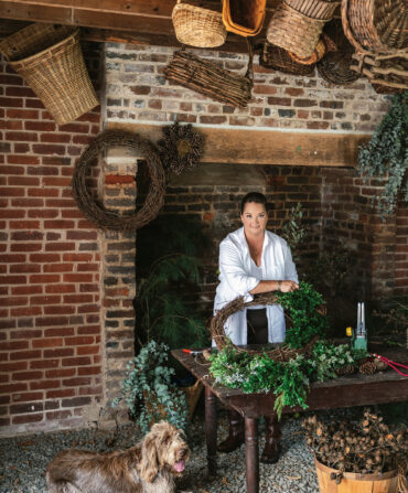 A woman stands behind a table with wreath-making supplies. There is a Spinone Italiano dog at her feet. A giant brick fireplace is behind her, and wicker baskets hang from the ceiling.
