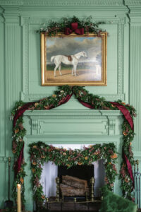 A grand garland of boxwood and olive with ribbon, dried oranges and lemons, hangs on a mint green mantle fireplace. A painting of a horse in a gold frame hangs above.