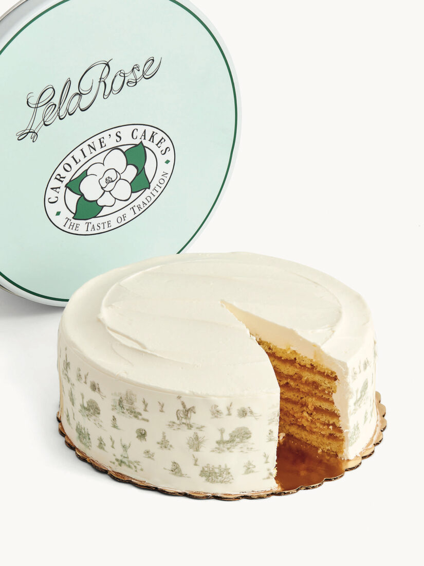 A white cake with a slice cut out to show layers of caramel and vanilla cake, and a blue-green tin behind it with the cake logo. The sides of the cake have a light green toile pattern printed on them