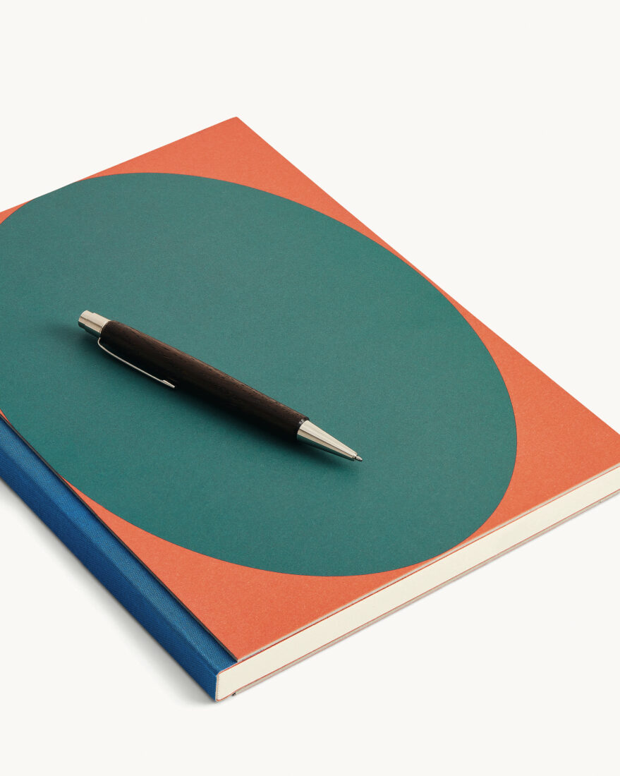 A orange notebook with a teal oval on the front and a deep blue spine. A black and silver pen rests on the notebook