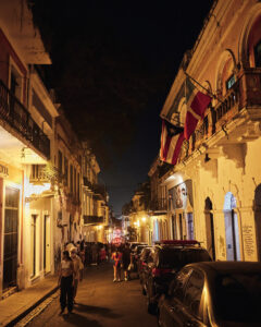 A lively nighttime streetscape in Old San Juan, with yellow lights and a Puerto Rican flag. People walk in the street.