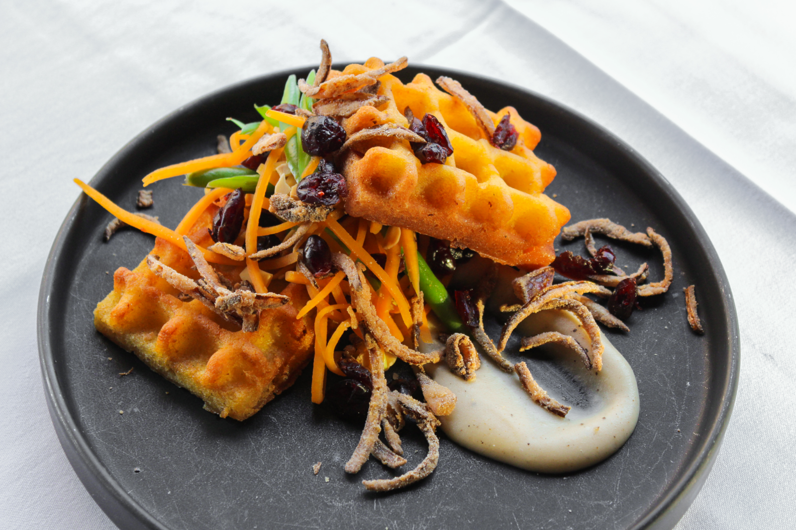 A white linen holding a round black plate, atop which sits two square orange waffles and a dab of a creamy beige sauce.