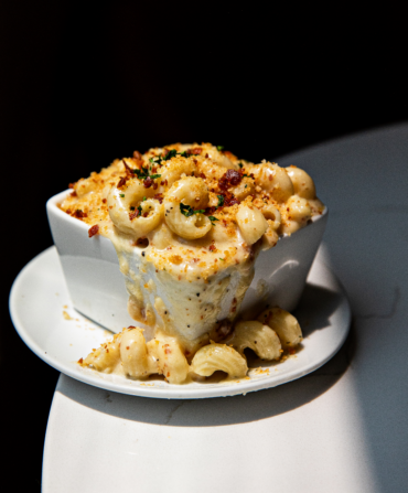 Macroni and cheese in a white square bowl atop a round white plate sits on a white table.