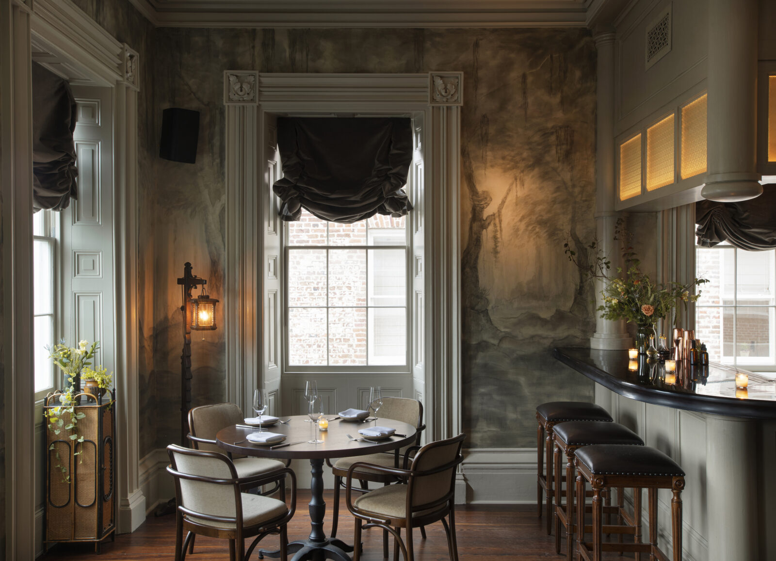 A dining room with a set table, painted wallpaper, and windows with draped curtains