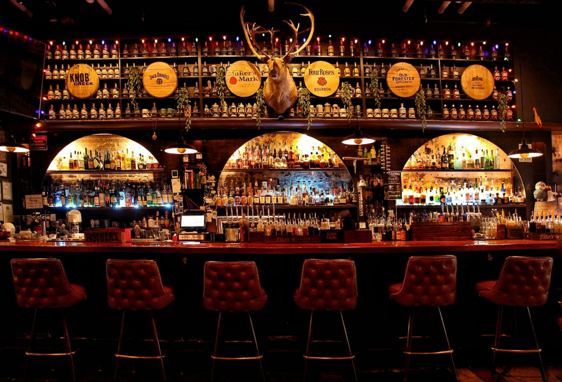 Burgundy colored quilted leather barstools line a dark wooden bar counter, behind which is a wall full of alcohol bottles, dim yet warm backlighting, and a mounted deer head.