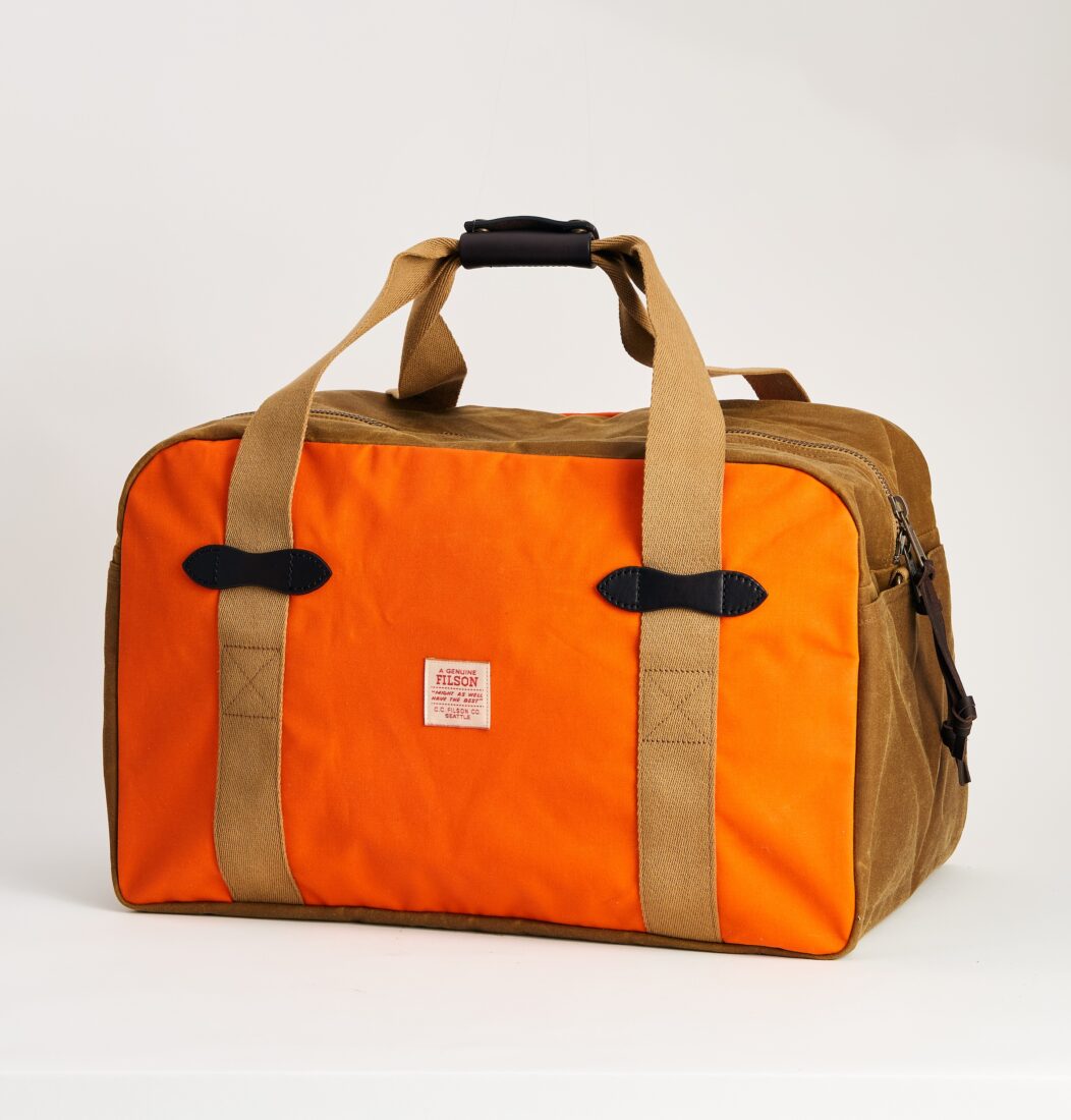 A bright orange duffle bag with brown straps and detailing; a hand is poised above it, ready to lift it