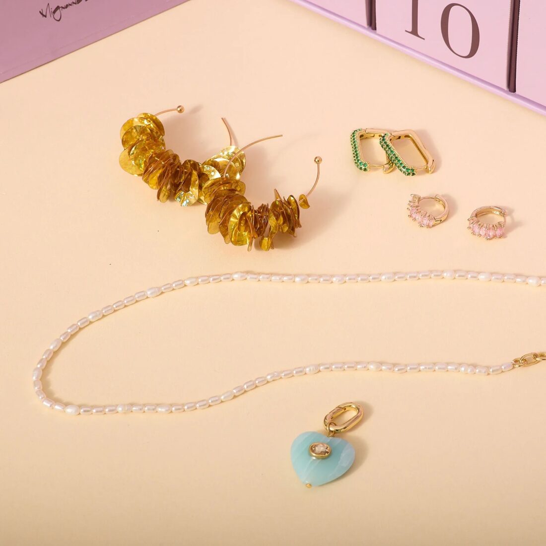 A pearl necklace, gold hoops, gold hoops with green jewels, gold hoops with pink jewels, and a blue heart-shaped pendant on a light peach background beneath a purple advent calendar box.