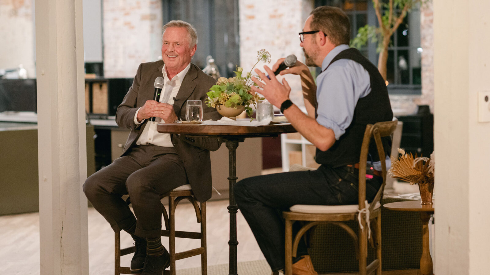 John Grisham and Jonathan Miles hold microphones while sitting next to each other at a table