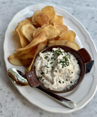 A white platter holds potato chips, a spoon, and a small dish with a creamy dip inside.