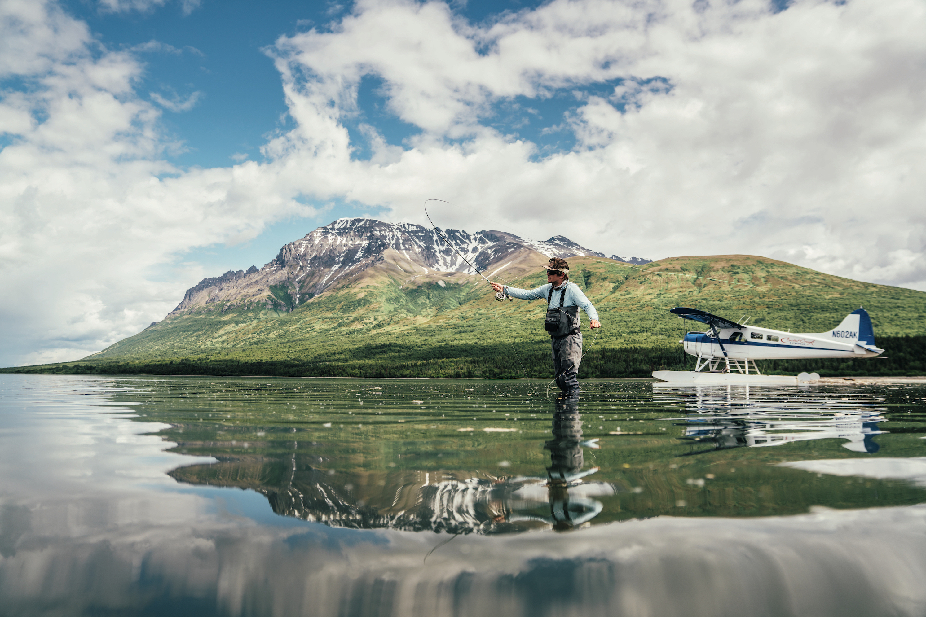 A man in waders casts a fly in water. Behind him there is a bush plane in the water and a mountain. The mountain landscape reflects in the water