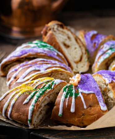 A king cake, sliced, with white icing drizzle, yellow, purple, and green spinkles, and a small pink plastic baby