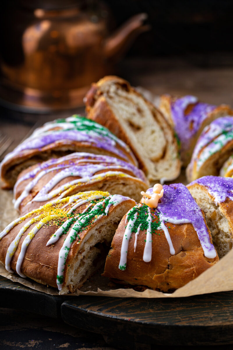 A king cake, sliced, with white icing drizzle, yellow, purple, and green spinkles, and a small pink plastic baby