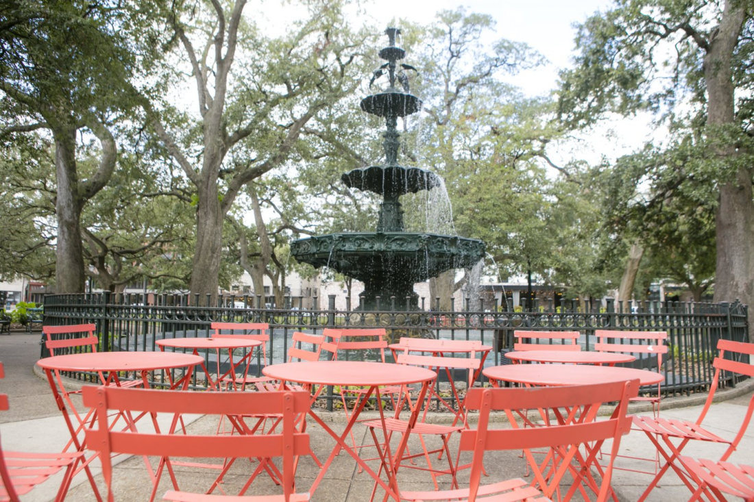 A tiered fountain is surrounded by a cluster of red tables and chairs, with threes in the background.