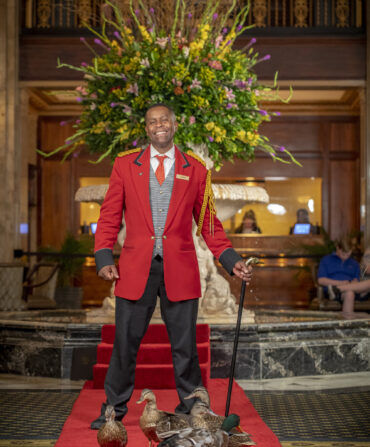 A man in a red jacket stands with a cane on a red carpet with several ducks at his feet