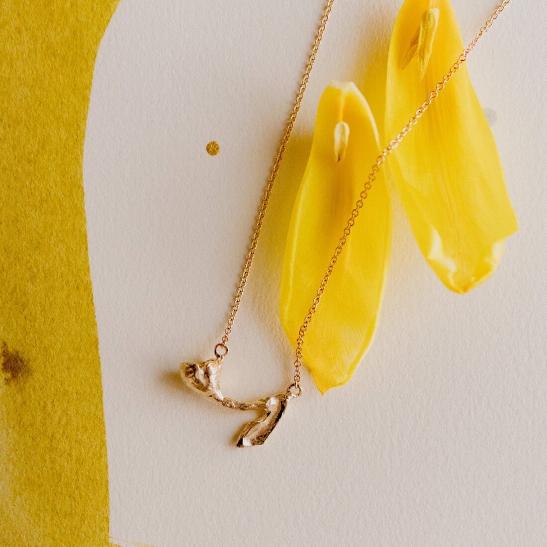 A necklace in gold with a tiny tulip pendant. It is on a background with yellow paint and two tulip petals that are yellow