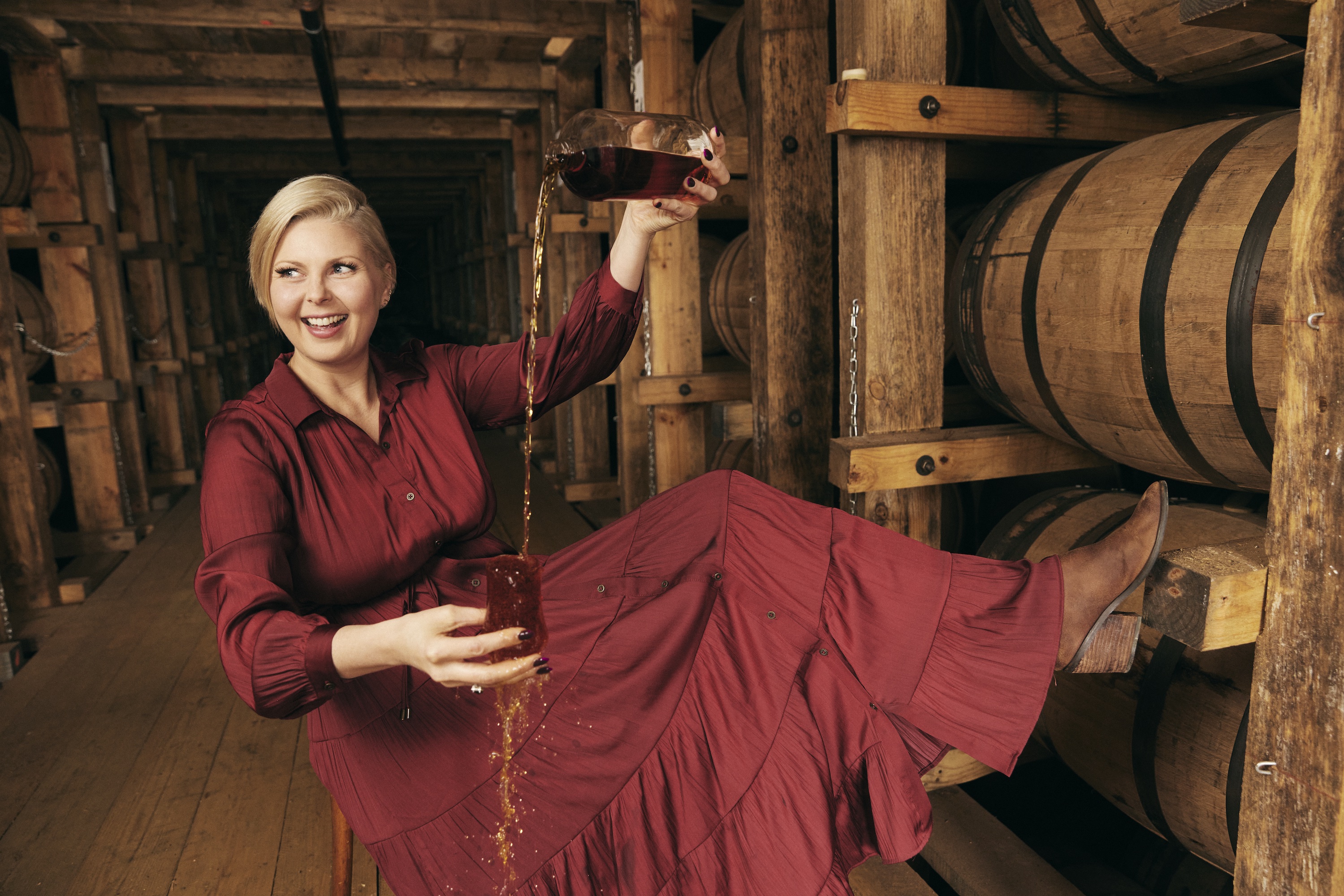 A woman holds a bottle of bourbon in a cask room. She pours it into a cup. She is wearing a red dress