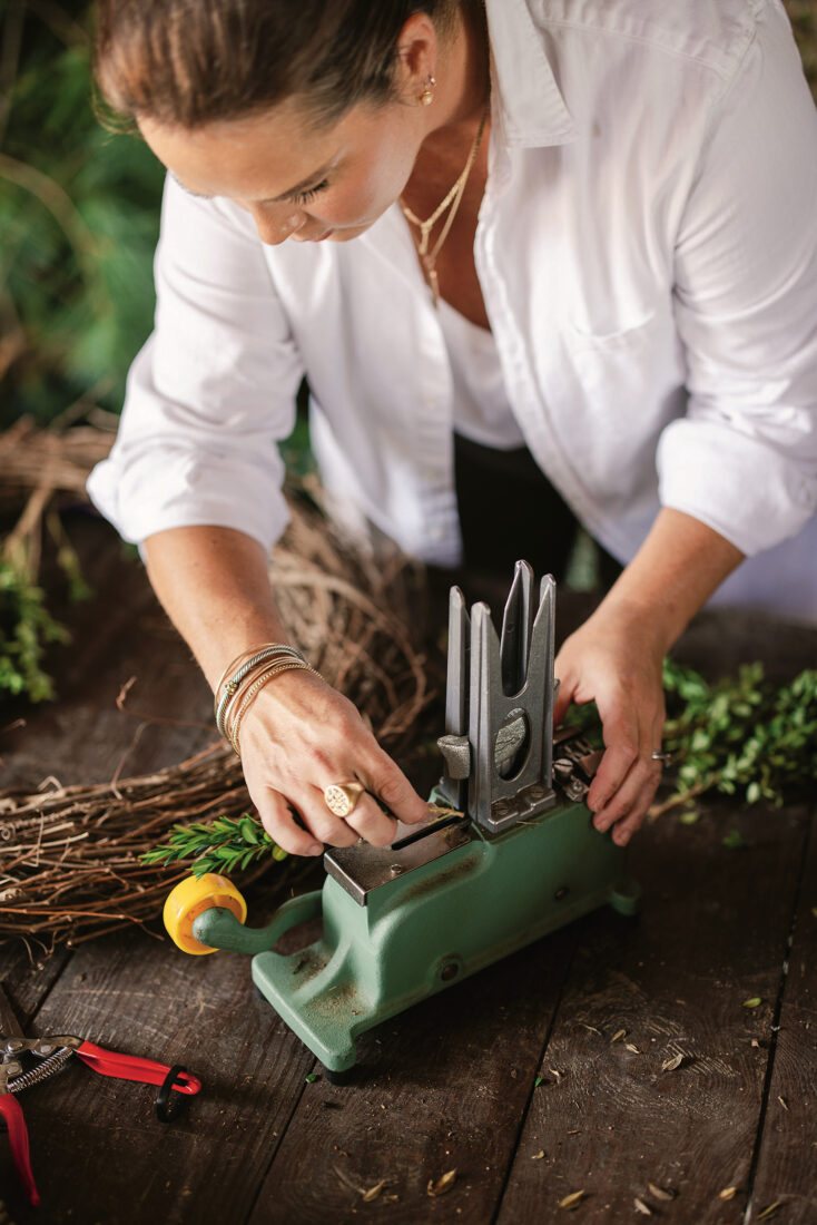 A woman presses a green vintage sewing machine on boxwood and cedar sprigs on a wood table.