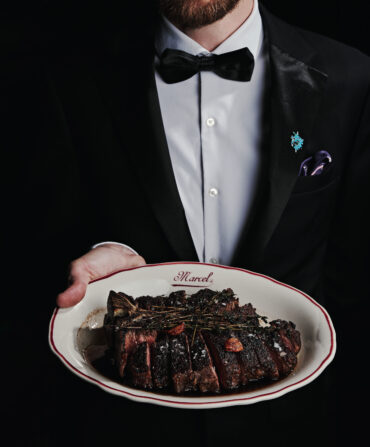 A tuxedoed waiter holds a plate with slices of steak