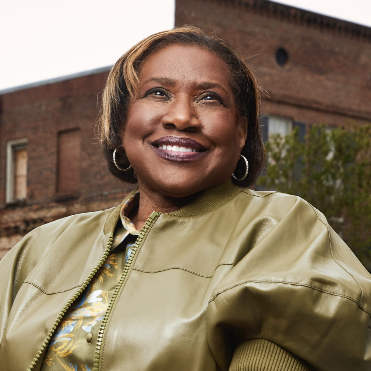 A smiling woman poses in front of a brick building and wears a green satin jacket, a green floral button down shirt, and hoop earrings.