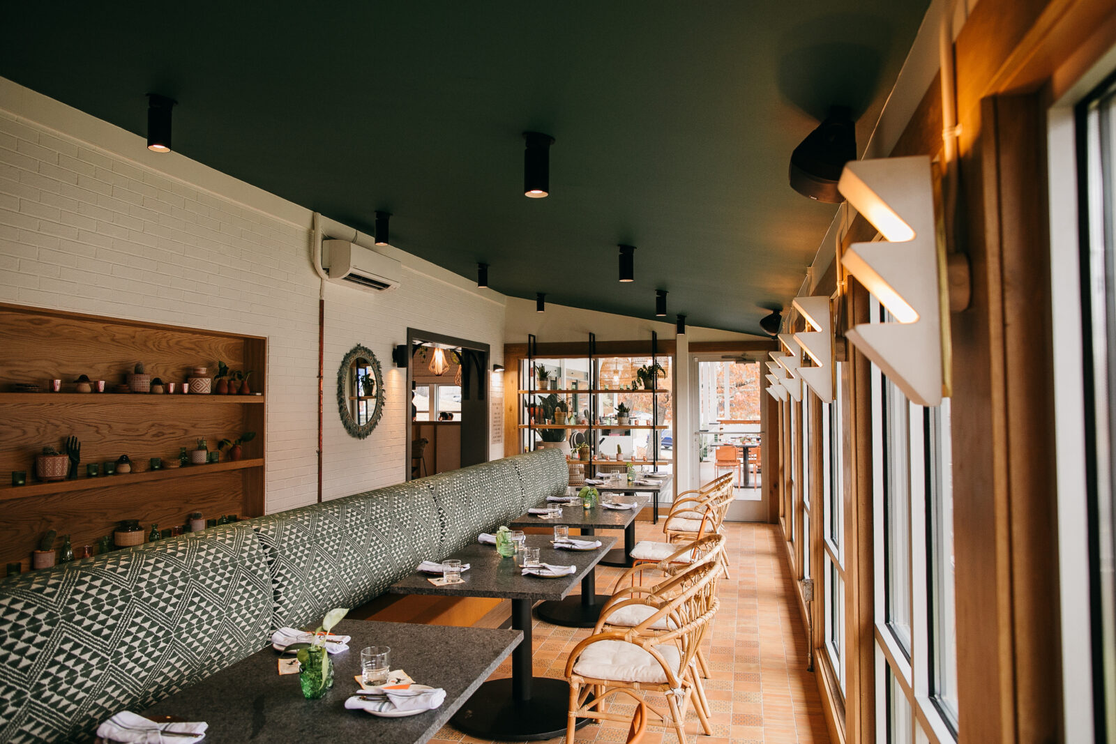 An airy porch dining area with wicker chairs, a black ceiling, a white brick wall, wood shelves, and booths patterned with green and white geometric fabric