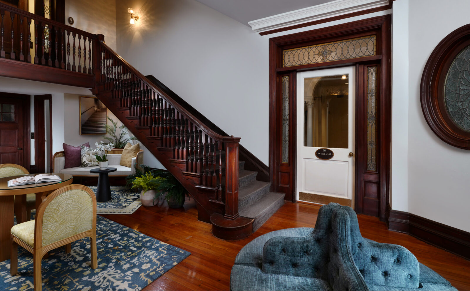 A lobby with dark brown stairs, blue seats and rugs, and a door with stained glass