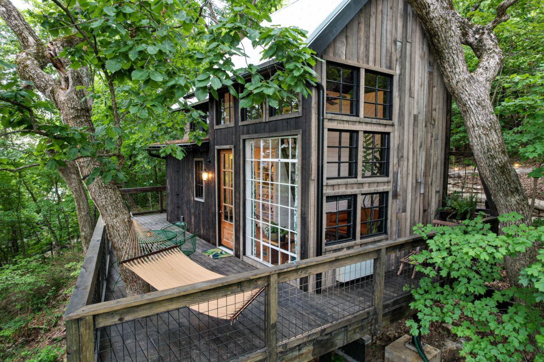 A large, grey wood treehouse with floor-to-ceiling windows is shrouded by trees and has a large deck with a hammock.