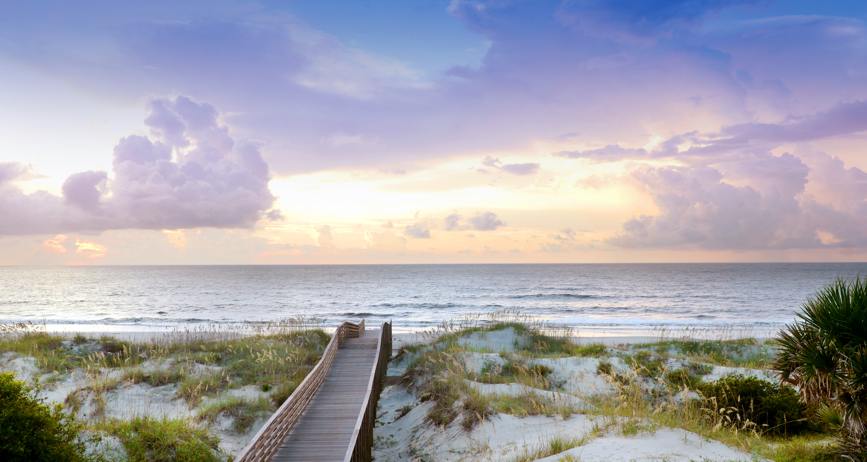 A beach and boardwalk; there is a purple and yellow sunrise