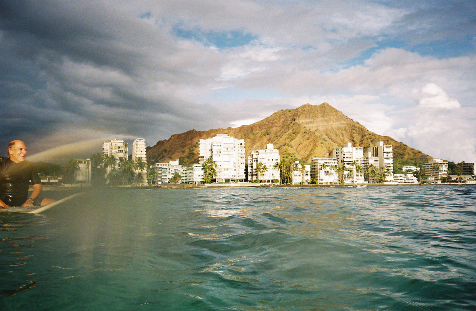 A man on a surfboard smiles in the water with a hilly town behind him