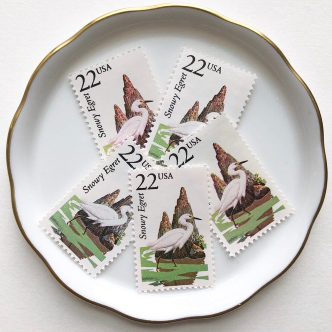 Five postage stamps with illustrations of a white egret. The stamps are on a white plate with a gold rim