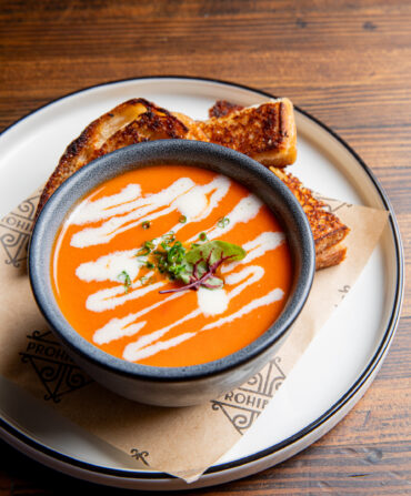 A bowl of tomato soup with a drizzle of white cream. Slices of grilled cheese are on a plate around the soup.