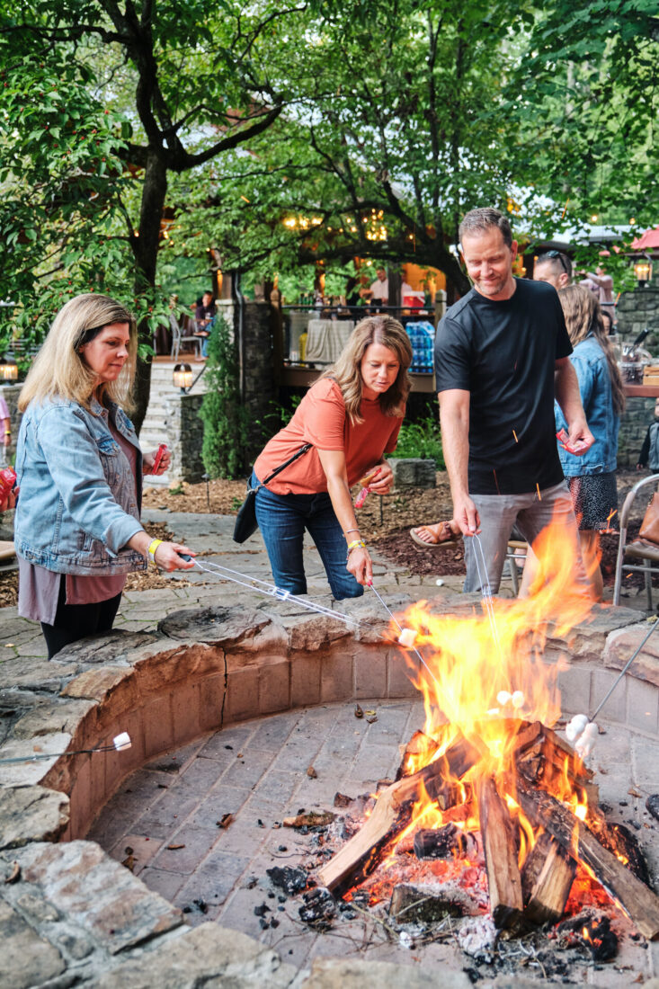 Two women and a man roast marshmallows over a fire pit