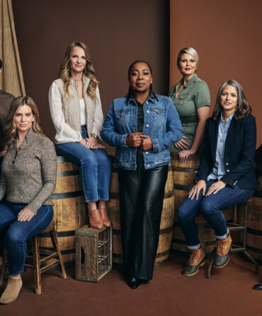 Seven women sit against wooden chairs, casks, and stools against a brown background.