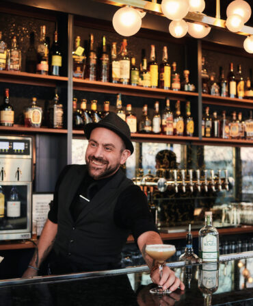 A man wearing a black vest and hat golds a whiskey sour at a black-topped bar. Walls of bottles are tucked into shelves behind him.