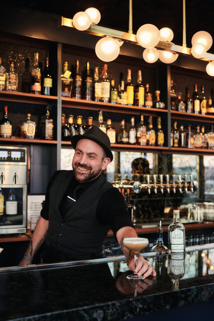A man wearing a black vest and hat golds a whiskey sour at a black-topped bar. Walls of bottles are tucked into shelves behind him.