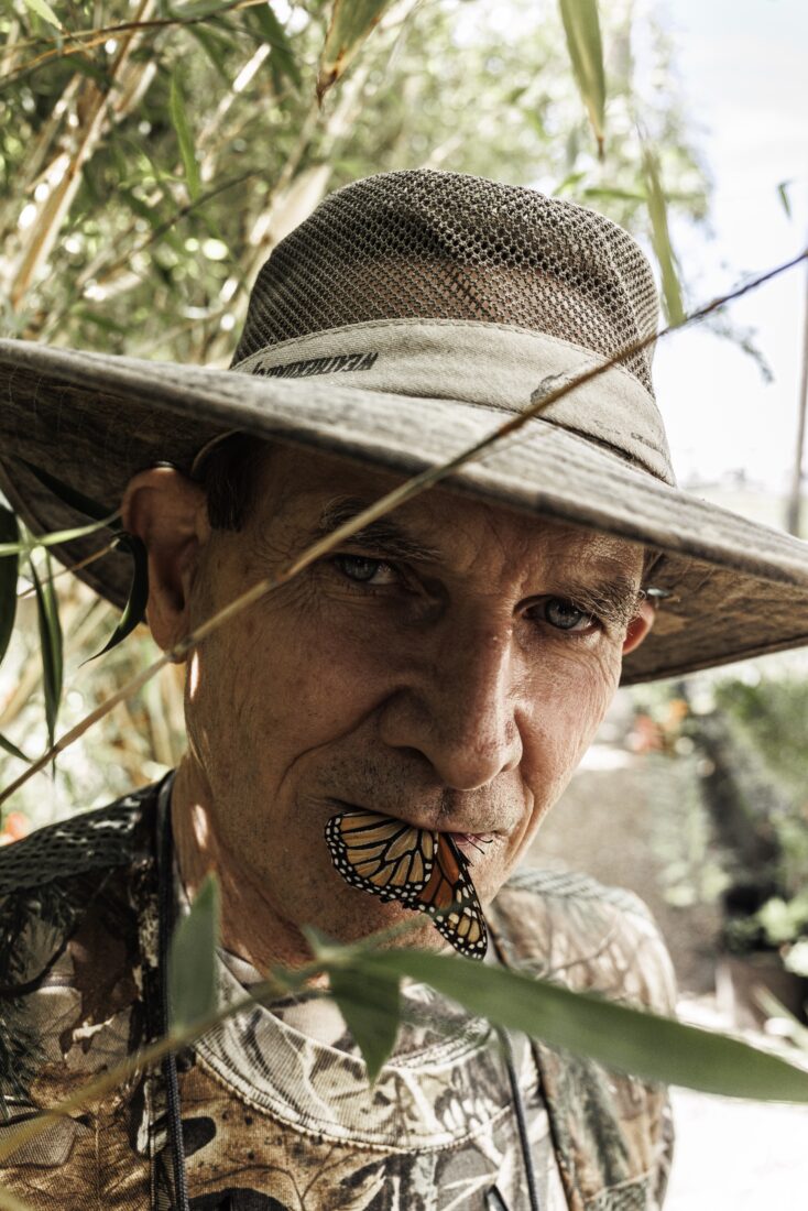 A man wearing camo and a hat holds a monarch butterfly gently in his mouth